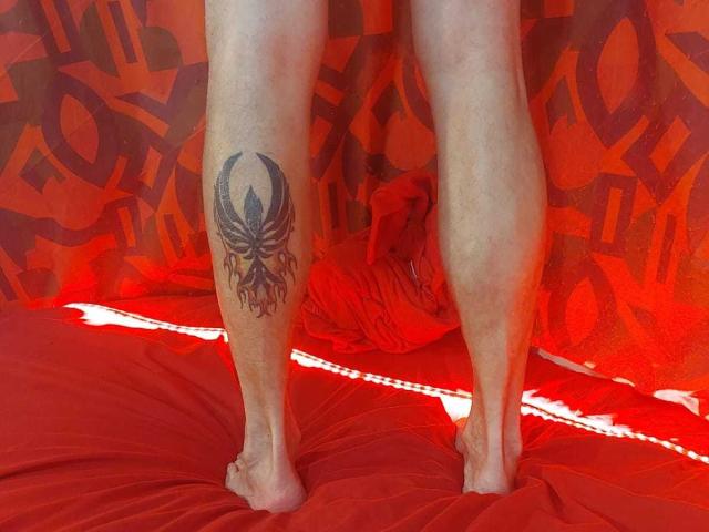 Michaell69's legs with a black and red tattoo of an octopus-like creature on the right calf, standing on a red bed with red curtains in the background.