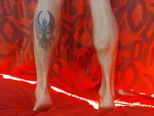 Michaell69's bare legs with a black tribal tattoo on the right thigh, standing on a red quilt, showing bare feet and muscular calves. Red cloth is bunched behind.