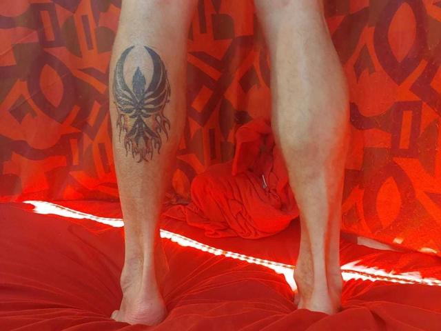 Michaell69's legs with a black skull tattoo on the right thigh, standing on a red bed with a red curtain in the background. Michaell69's feet are bare.