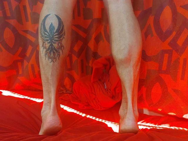 Michaell69's legs with a black tattoo of a detailed skull on the right calf, standing on a red, textured surface, barefoot, with a red cloth on the floor.