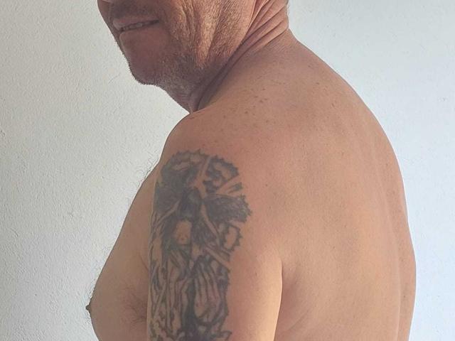 Michaell69's shirtless upper body shows a tattoo on his right shoulder of a dark, detailed, tree-like design with a face in the center. His face is partially visible.
