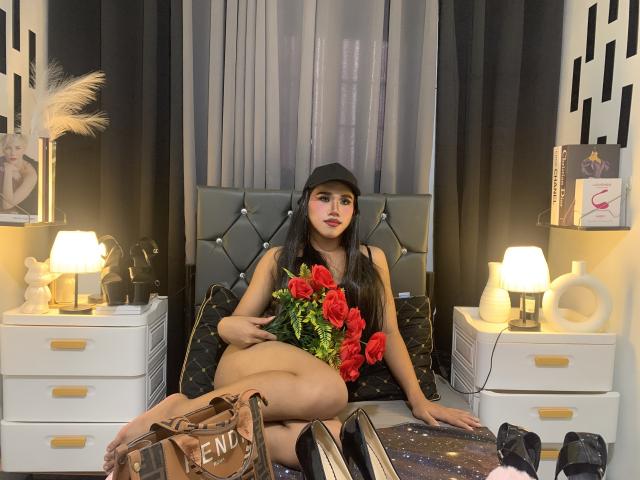 YeshaSebastian, wearing a black cap, sitting on a bed with red roses, black lingerie, and a small ass, surrounded by white drawers, black heels, and a beige bag.