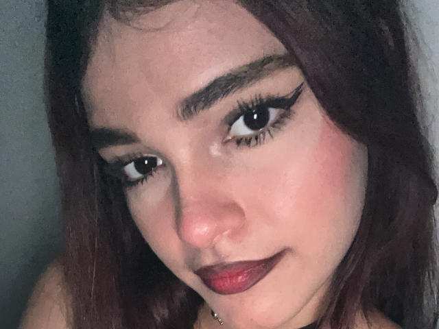 CameronBrown's face with dark red lipstick, thick black eyeliner, and brown wavy hair, looking down with a slight smile, showing a nose ring and subtle makeup.