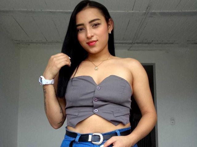 MaraLewis with long black hair, gray strapless top, blue jeans, white watch, small tits, tan skin, red lipstick, tattoo on left arm, smiling, holding hair.