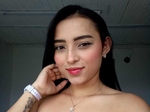 MaraLewis has long black hair, pink lipstick, and a small nose ring. She's wearing a white wristband, earrings, and a necklace with a butterfly pendant. Her hand's on her cheek.