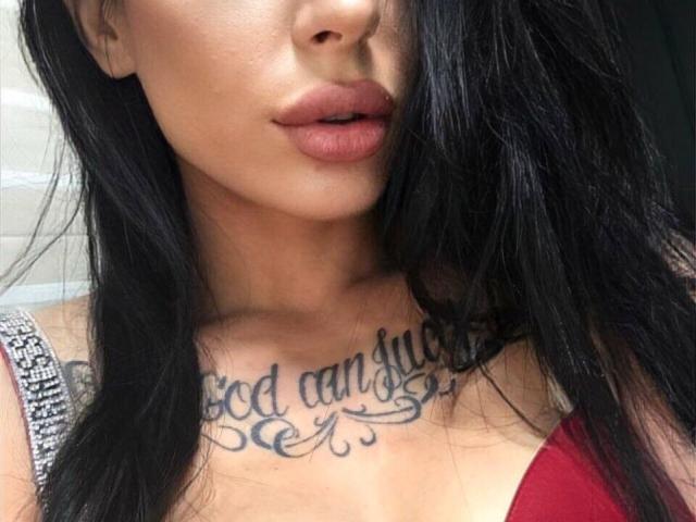 PamelaMoon's face with black hair, red lipstick, and a tattoo on her chest reading "God can" in cursive. Her tits are visible in a red top.