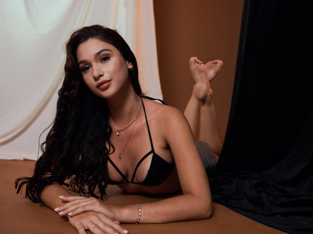 BellaBee with long black hair, lying on brown floor, wearing black bikini top, showing small tits, legs up, brown skin, confident smile, necklace, bracelet, bare feet.