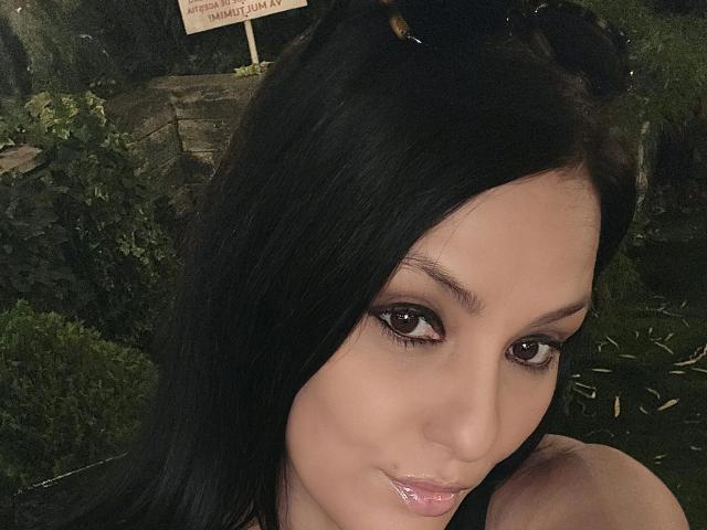 JoliDanielle with black hair, brown eyes, and shiny pink lips, smiling at the camera, showing her cute face and smooth skin. Her boobs are slightly visible, but not the focus.