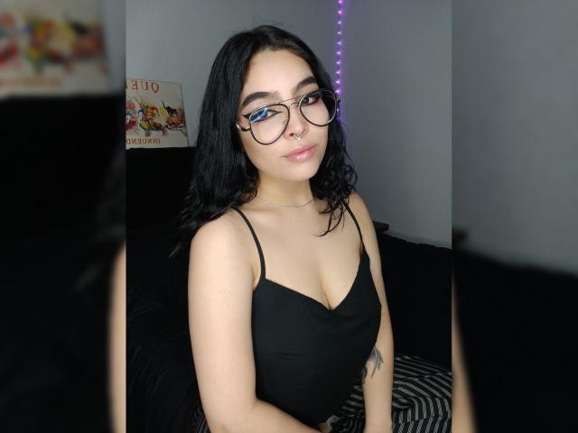 MeghanRider with dark hair, glasses, nose ring, black tank top showing her tits, choker, tattoo on right arm, sitting on black bed, purple string lights in background.