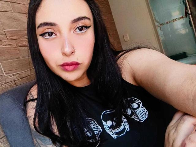 AmberGoddesss with long black hair, dark eyeliner, pink lipstick, black skull shirt, nose piercing, and calm expression, sitting with arms crossed.