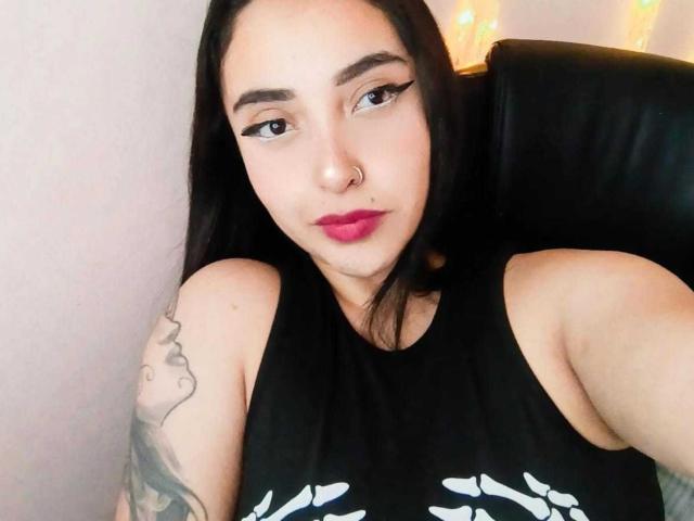 AmberGoddesss with long black hair, nose ring, and red lipstick, wearing a black sleeveless top, sitting in a black leather chair, showing a tattoo on her left arm.