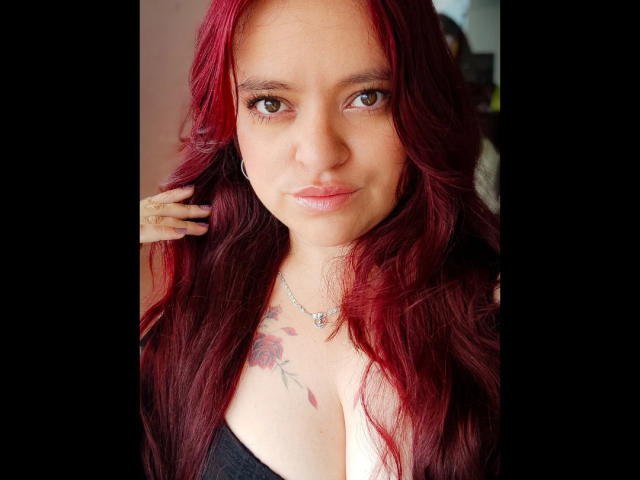 ElizabethFire with red hair, brown eyes, and a tattoo on her chest, wearing a black top, showing her tits, has a necklace, and is looking at the camera.