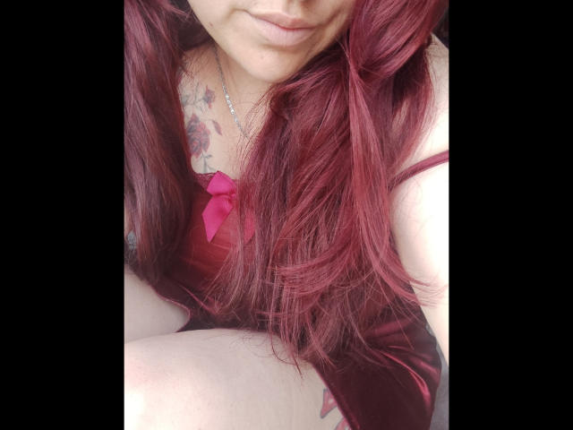 Elizabeth: Cabelos ruivos, um laço rosa no vestido e uma tatuagem no peito, mostrando os seios e um sorriso discreto. Seus joelhos são visíveis em primeiro plano..