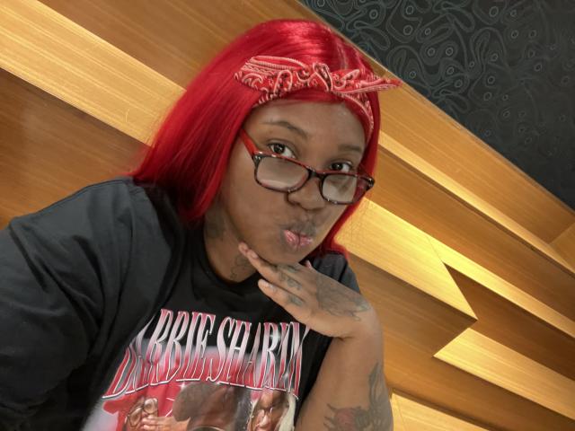 Shabrim with red hair, glasses, and bandana, pouting with finger on lips, wearing a black Britney Spears shirt, showing tattoos on hands and arms.