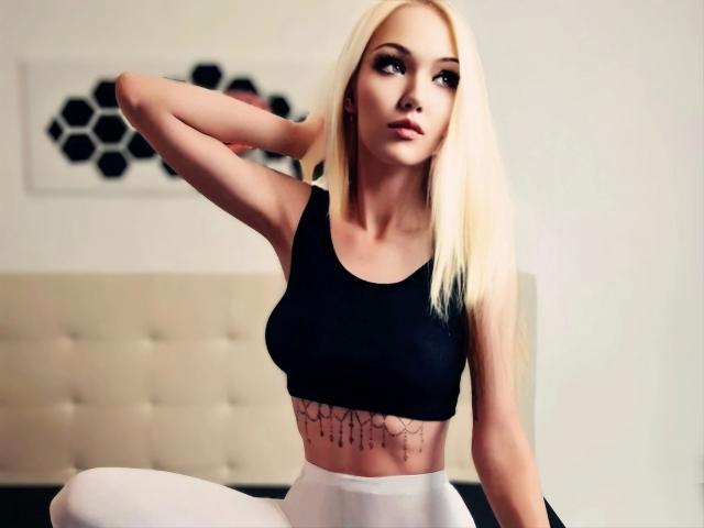 Ginacolada-hot with blonde hair, wearing a black sports bra, white pants, showing a tattoo under her tits, arm raised, looking away.