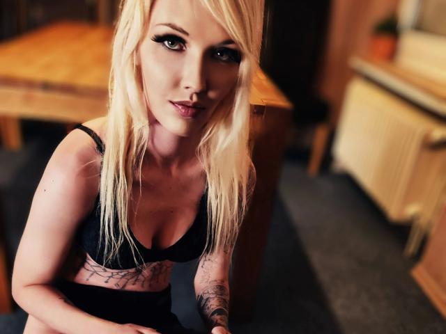 Ginacolada-hot with blonde hair, black lingerie, and tattoos, looking up with a slight smile, showing her tits and a tattoo on her right arm. Blurred indoor background.