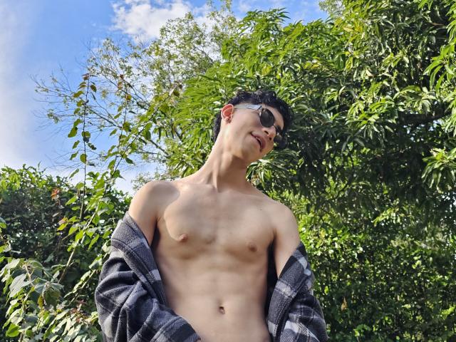 NovaWheeler, shirtless with a plaid jacket hanging off, wearing sunglasses, has a slim, toned, and hairy chest. He's standing in a green, leafy outdoor setting.