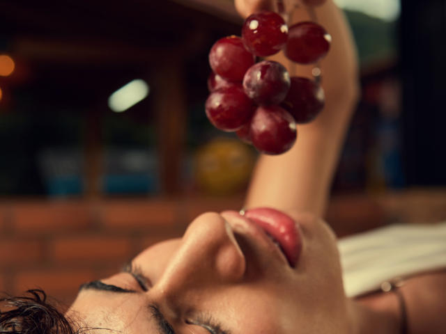 NovaWheeler's face is upside down, tongue out, holding a cluster of red grapes near her mouth. Her eyes are closed, and her nose is slightly upturned.