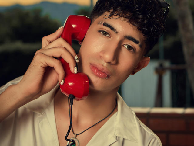 NovaWheeler with curly dark hair, tan skin, and pouty pink lips, holding a red phone to his ear, wearing a white shirt, and a black necklace.