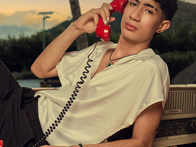 NovaWheeler with short brown hair, wearing a white shirt and black pants, holding a red coiled phone to his ear, looking at the camera with a slight pout.