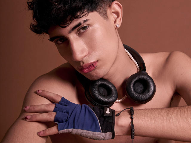 NovaWheeler's shirtless, wearing black headphones, a pearl necklace, and a blue fingerless glove. He has curly black hair, a nose ring, and a brown background. 