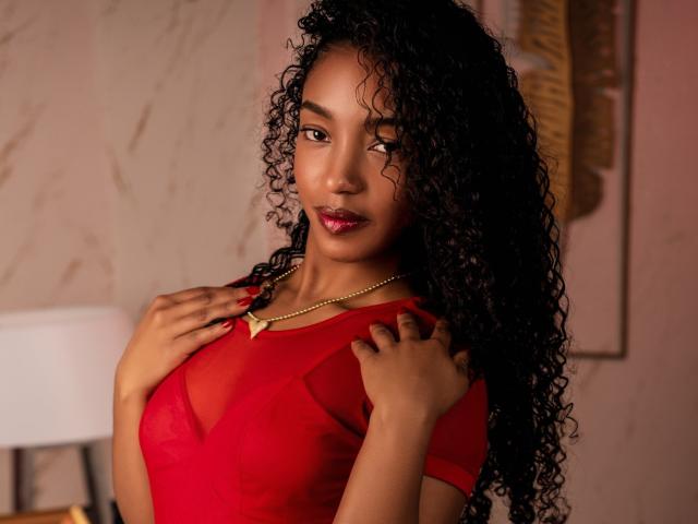 AlonnaGonzales, con cabello negro rizado, vestida con una blusa roja transparente, con las manos en el pecho, lápiz labial rojo, collar dorado, mirando a la cámara, con una expresión segura, atractiva, piel oscura, iluminación cálida..
