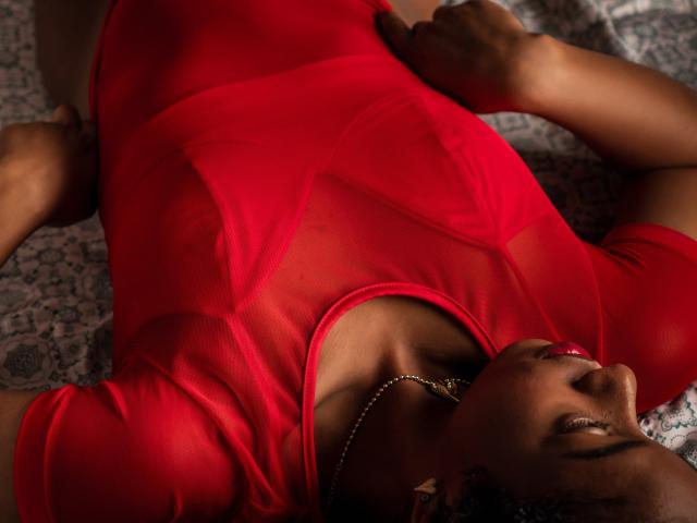 AlonnaGonzales is lying down in a sheer red dress, exposing her tits, with her hands on her ass and a gold necklace, looking up with red lipstick.