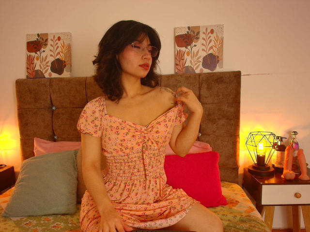 SeleneLeon with dark wavy hair, red lipstick, and glasses, wearing a pink floral dress, sitting on a bed, slightly pulling up the dress to show her tits.
