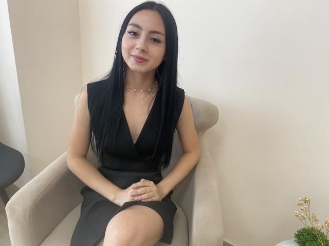 MiyaStiverts with long black hair, wearing a black dress with a deep V, sitting in a beige chair, hands on lap, subtle smile, necklace, light skin, soft makeup.