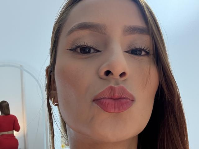 MyaSpecter's face with pouty pink lips, brown eyebrows, long eyelashes, and straight brown hair. Red outfit reflected in mirror on left. Gold hoop earrings. Soft natural light.