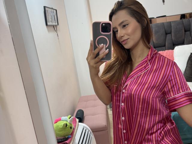 MyaSpecter with long brown hair, wearing pink and purple striped pajama top, holding pink phone, taking selfie in mirror, smiling, pink cushion, green toy in background.