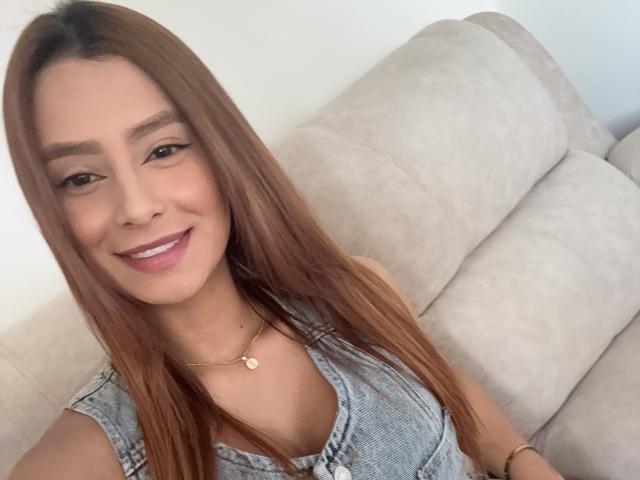 MyaSpecter has long brown hair, smiling with pink lipstick, wearing a denim top, and a gold necklace with a heart pendant, sitting on a beige couch.