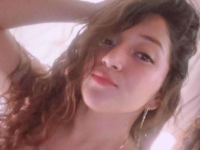 NicoleMartins with wavy brown hair, wearing hoop earrings, has soft pink lips and a slight smile, looking at the camera with her hand in her hair.