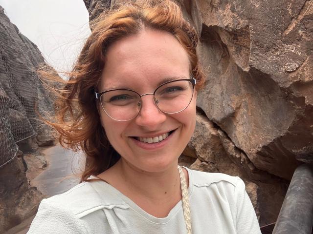 SophieShy-hot has red curly hair, glasses, and a white textured shirt, smiling with visible teeth, standing against rocky brown cliffs, showing a cute, natural, and happy face.
