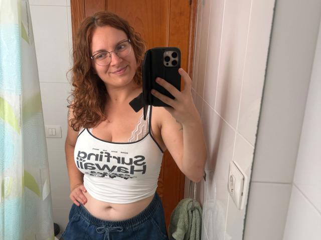 SophieShy-hot with curly red hair, glasses, white tank top showing her boobs, and blue pants, holding a black phone, smiling in a bathroom mirror selfie.