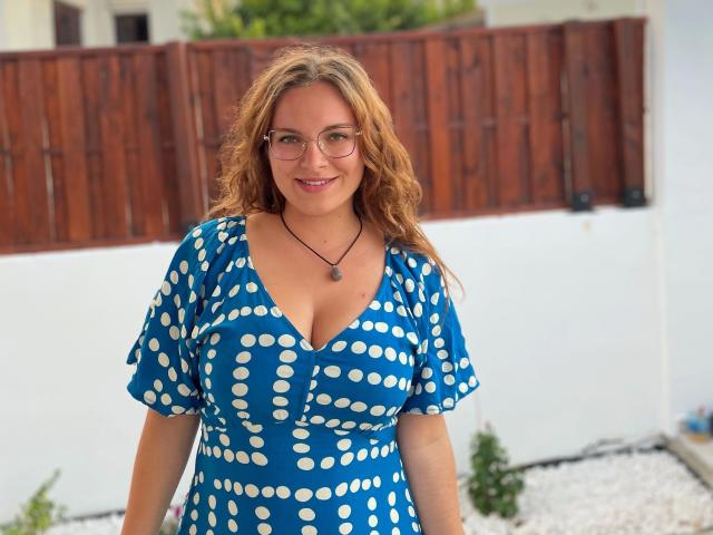 SophieShy-hot with curly blonde hair, glasses, blue polka dot dress, and cleavage, smiling in a backyard. She's wearing a black necklace with a purple stone.