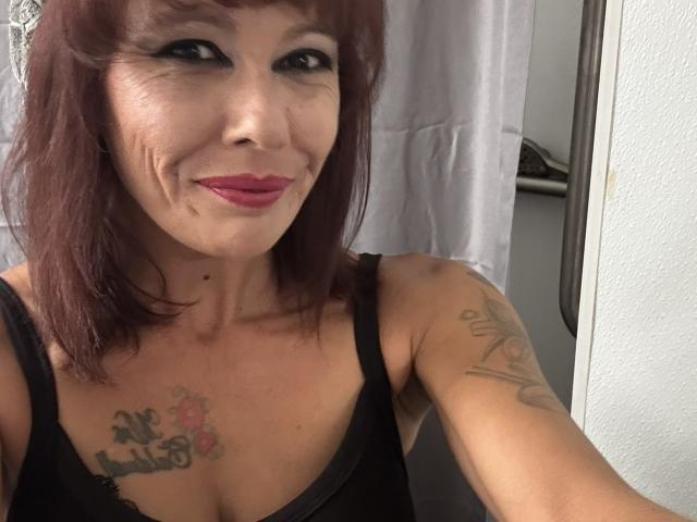 LizzieStar69 with red hair, black tank top, red lipstick, tattoos on chest and arm, smiling, holding up phone for selfie. Boobs visible under tank top.