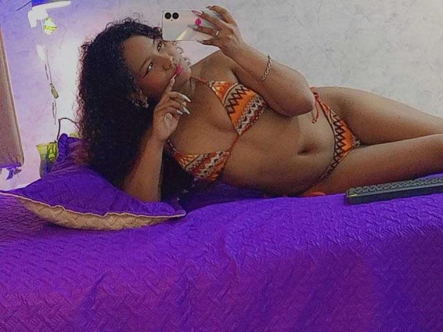 EvaBennet, lying on a bed with purple sheets, wearing an orange and yellow zigzag bikini, taking a selfie with a phone, showing her tits and smiling.