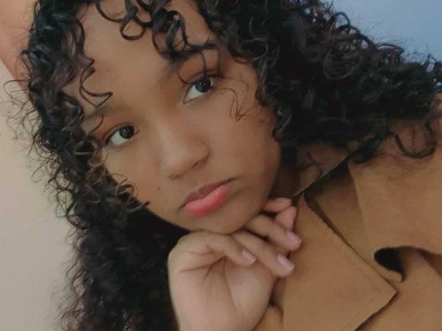 EvaBennet has curly black hair, brown skin, and pink lips, resting her chin on her hand with a slightly pouty expression, wearing a brown coat.
