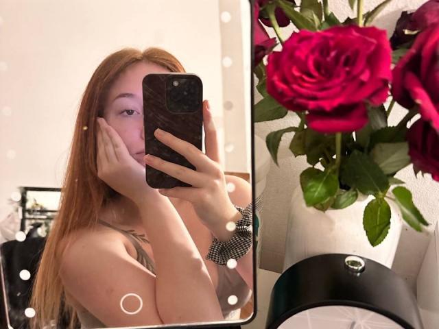 Barberry with long red hair, hands on cheeks, taking a mirror selfie, showing her bare shoulders and a black phone. Red roses in the foreground.