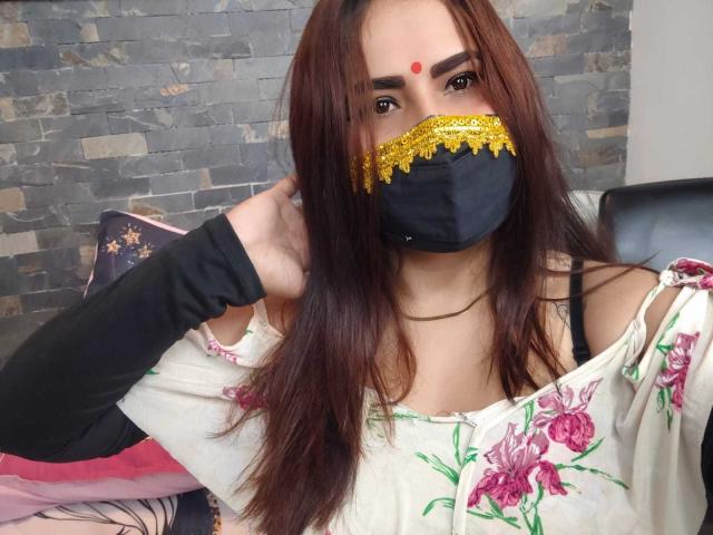 Dalia Dubey, con cabello castaño, un bindi rojo, una máscara negra con encaje amarillo, una blusa con estampado floral, mangas negras y un collar dorado, mirando a la cámara con una mano en la cabeza..