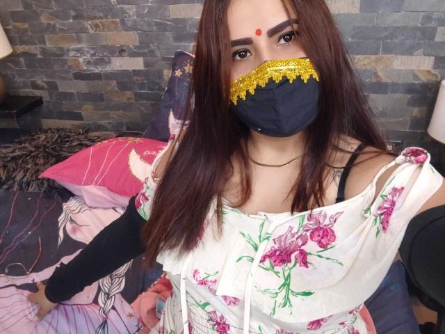 Dalia Dubey, con cabello largo y castaño, vestida con un vestido blanco con estampado floral, una máscara negra con detalles amarillos y un bindi rojo, mostrando ligeramente sus senos a través del vestido, sobre una cama..