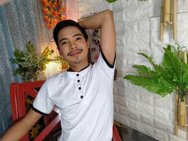 MasterManiacXx with short black hair, a small mustache, and a white shirt with black buttons, arms raised, smiling, in a room with green plants and a white brick wall.