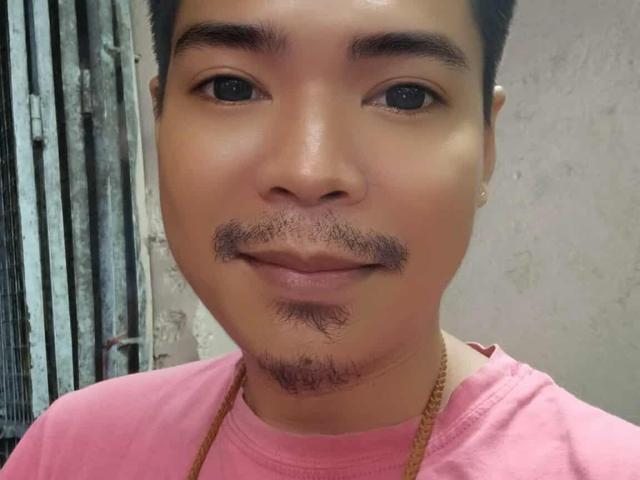 MasterManiacXx with short black hair, light brown skin, small goatee, and mustache, wearing a pink shirt and gold necklace, smiling at the camera.