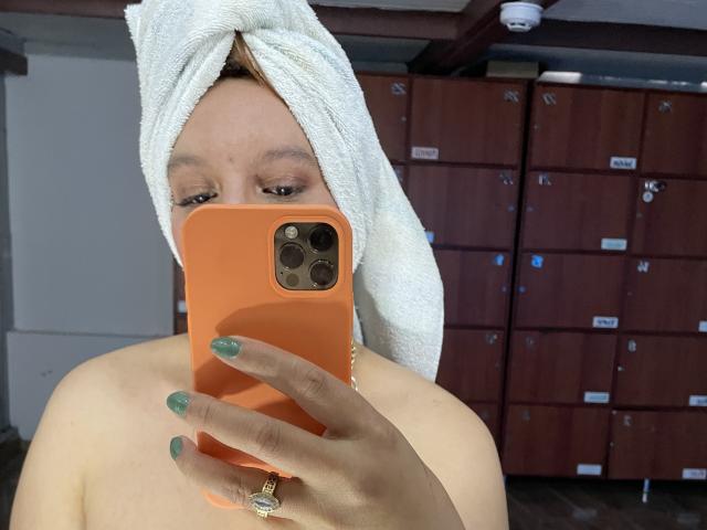 RoxieJackson with a towel on her head, holding an orange phone, showing green nails, and a gold ring, taking a selfie in a locker room.