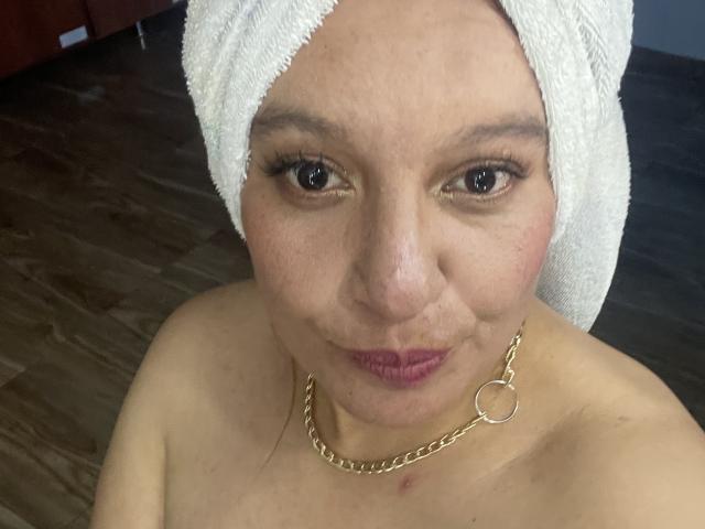 RoxieJackson with a white towel on her head, wearing a gold chain necklace, has brown eyes, light skin, and pink lipstick, showing a small red mark on her chest.
