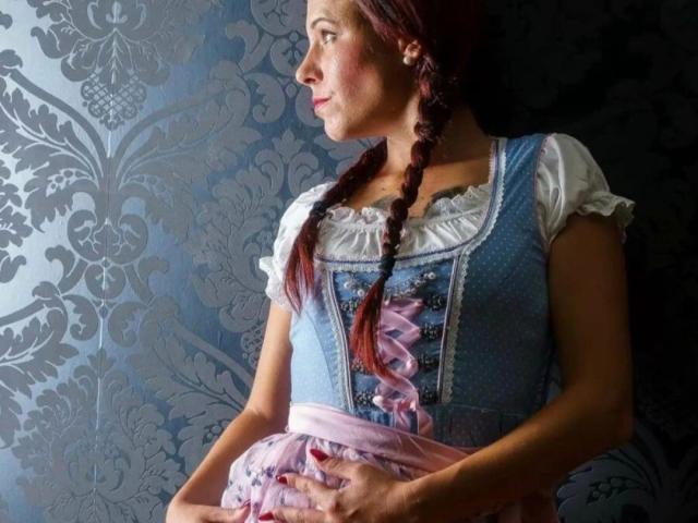 SexyAnnabell38-hot with braided red hair, wearing a blue dress with pink ribbons, white shirt, holding her ass, looking to the side, against a dark, floral-patterned background.