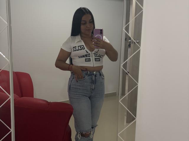HikariChloe with long black hair, white crop top showing her small tits, and high-waisted blue jeans, standing in front of a mirror, holding a pink phone.