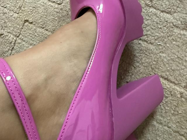 Margos Fuß in einem glänzenden, pinken High Heel-Schuh mit Blockabsatz, der ihren Knöchel und das Schuhband zeigt. Teppichboden im Hintergrund..