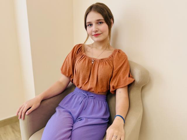RebeccaChamb sitting in a beige chair, wearing an orange short-sleeve top and purple high-waisted pants, smiling slightly, brown hair in a loose ponytail, blue bracelet on right wrist.